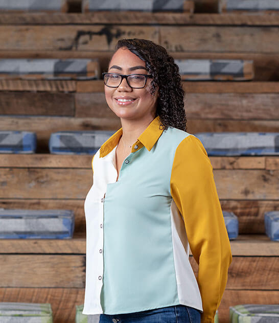 Woman in a white, seafoam, and mustard shirt with glasses standing in front of wooden stairs